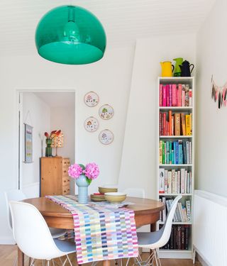 White dining area with green glass pendant light and colour coded books on bookshelf