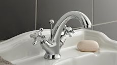 a silver tap, one of the best bathroom taps, on a white bathroom sink with a bar of soap next to it, against a grey tiled wall