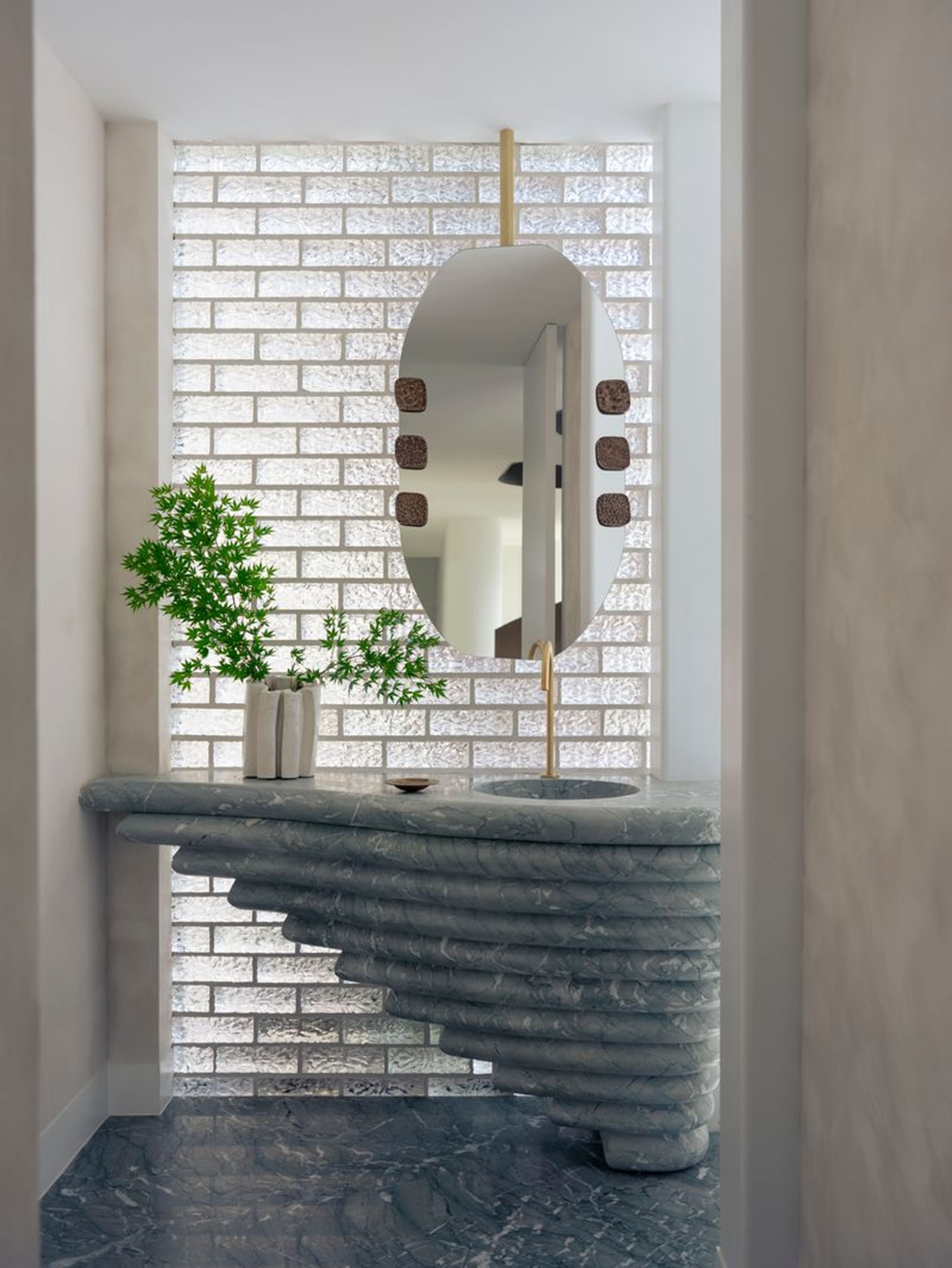 a bathroom with a glass block wall, sculptural marble vanity, suspended bathroom mirror and vase with a branch in it