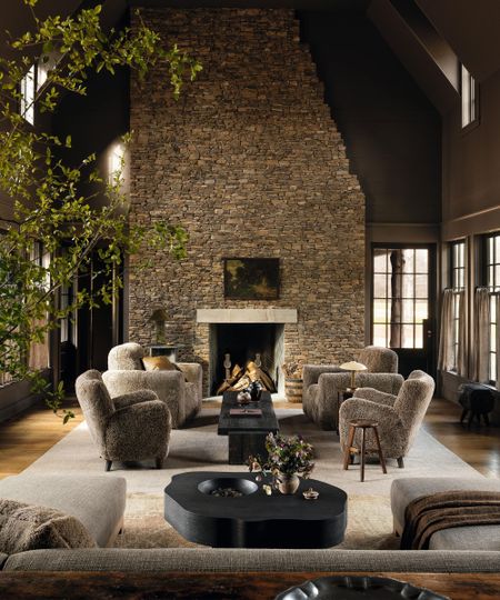 a large great room in a farmhouse home with a towering stone fireplace and shearling armchairs circled around a black rustic coffee table