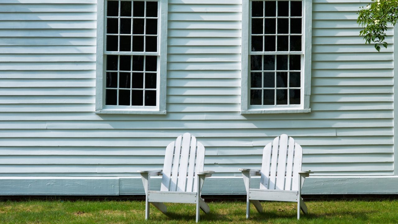 Light blue siding on a close up of exterior of a house. There is green lawn and two white Adirondack chairs