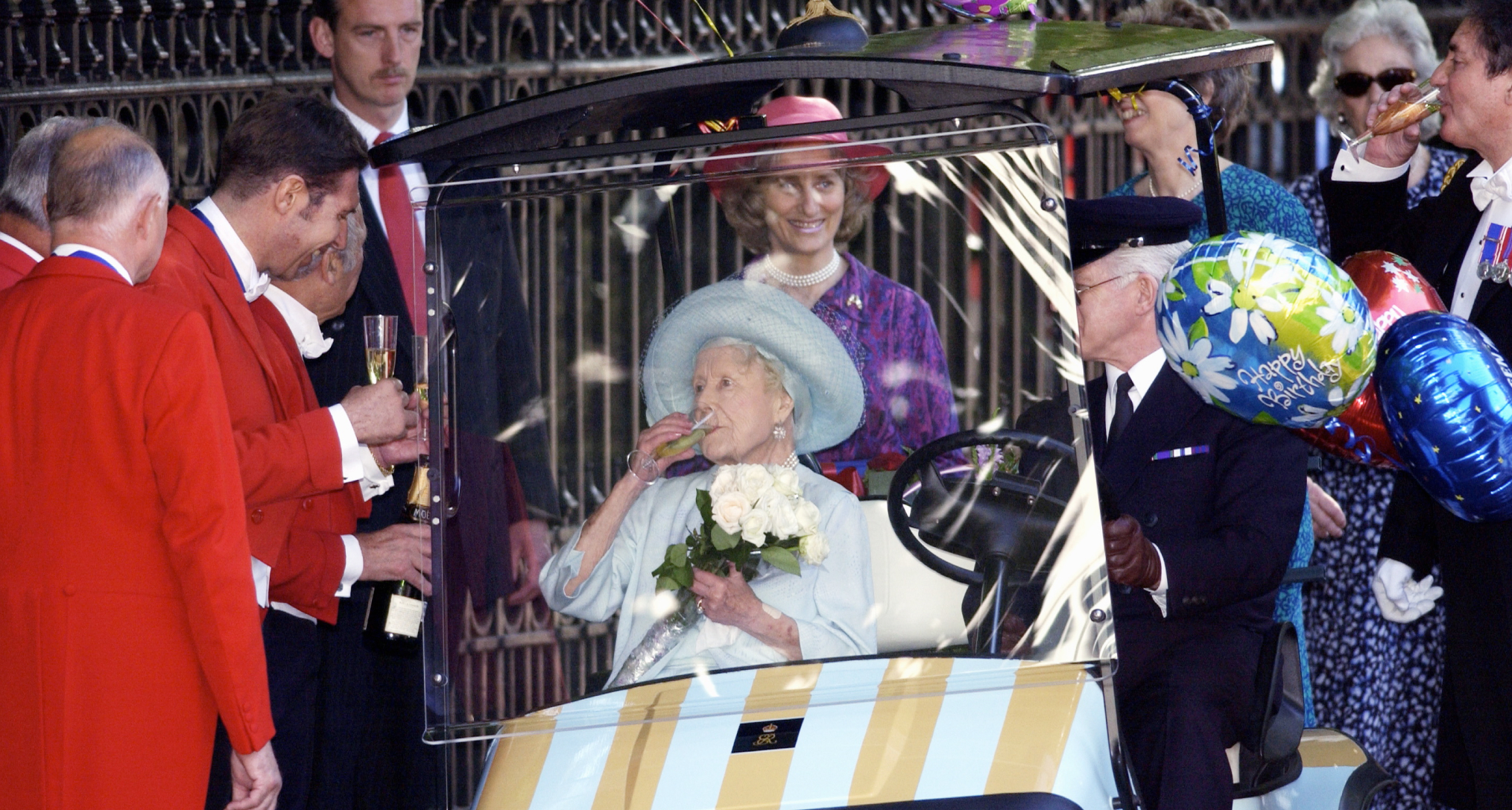 The Queen Mother drinking champagne while being driven around in a buggy as staff members drink champagne next to her