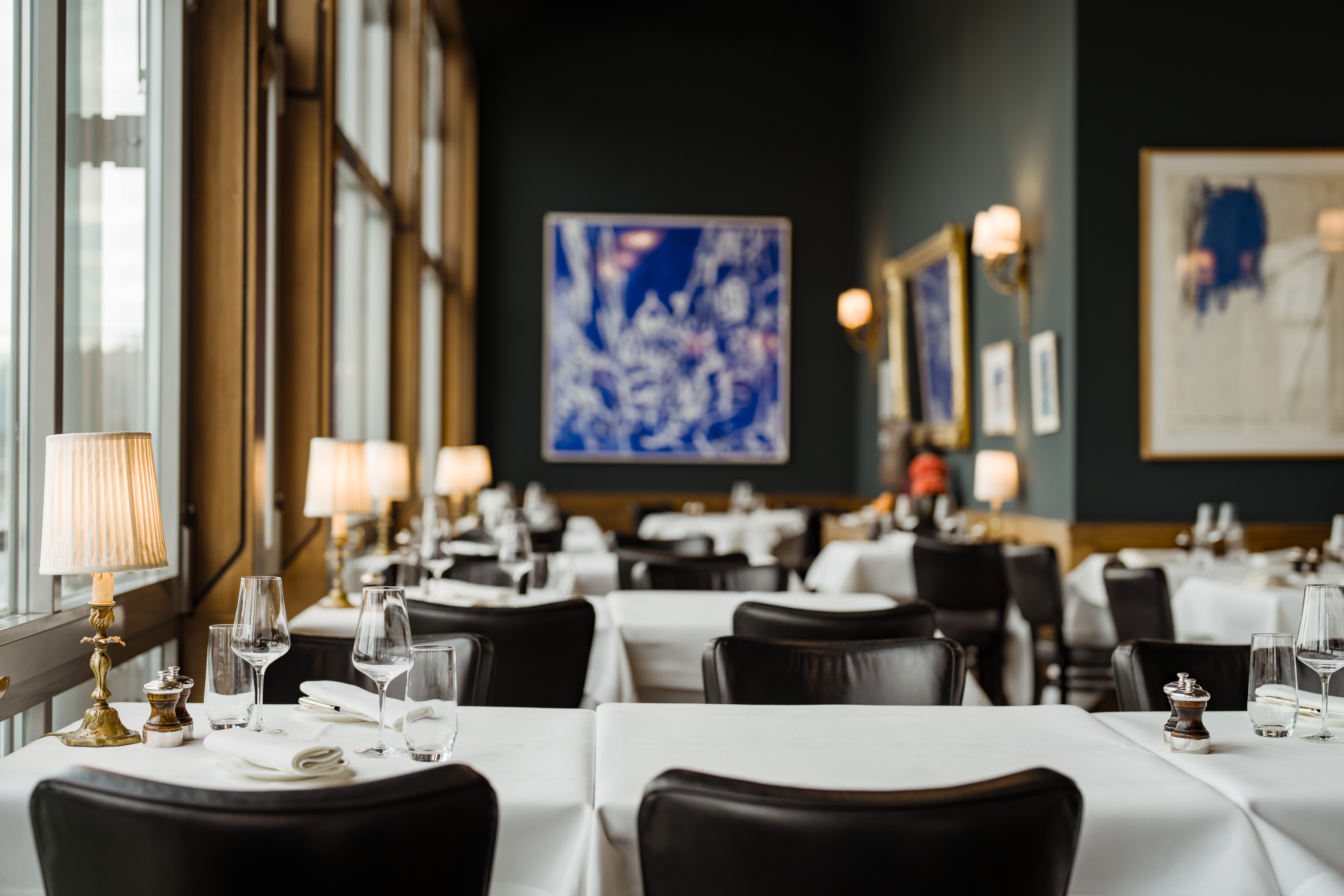 The Brasserie inside Les Trois Rois, a restaurant with black leather chairs and crisp white tablecloths, and small gold lamps and wine glasses on each table, the walls are painted black.