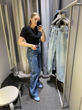 Woman takes photo in dresing room mirror, wears black t-shirt, blue jeans, blue trainers