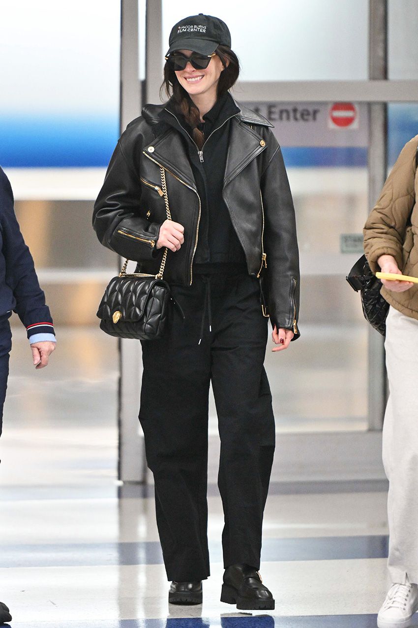 Anne Hathaway at the airport in NYC wearing a black leather jacket, half-zip pullover, black pants, black chunky shoes, a black quilted bag, and baseball cap
