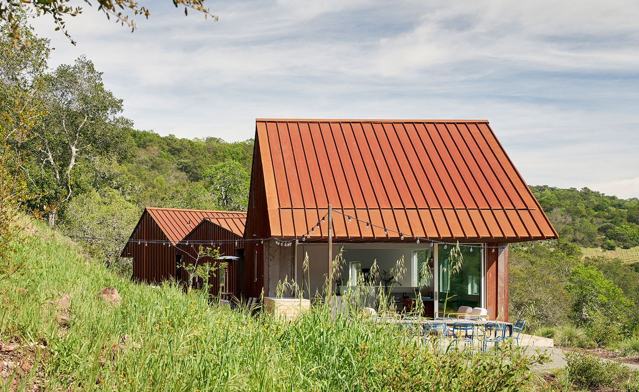 Exterior view of Triple Barn house by Mork-Ulnes Architects featuring three Corten steel roofs during the day. The house is surrounded by greenery and has strings of lights outside