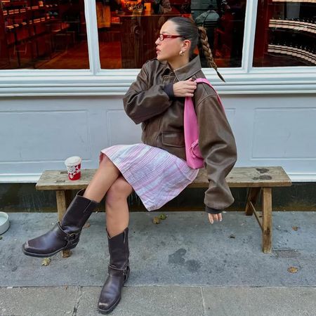 a woman wearing a brown leather jacket, pink shoulder bag, pink striped skirt, and brown boots