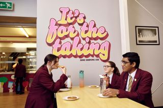 School children eating lunch next to a wall with the quote 'It's for the taking' on it