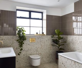 A neutral bathroom with stone tiles onthe lower half of walls, a wall mounted toilet, and square tub