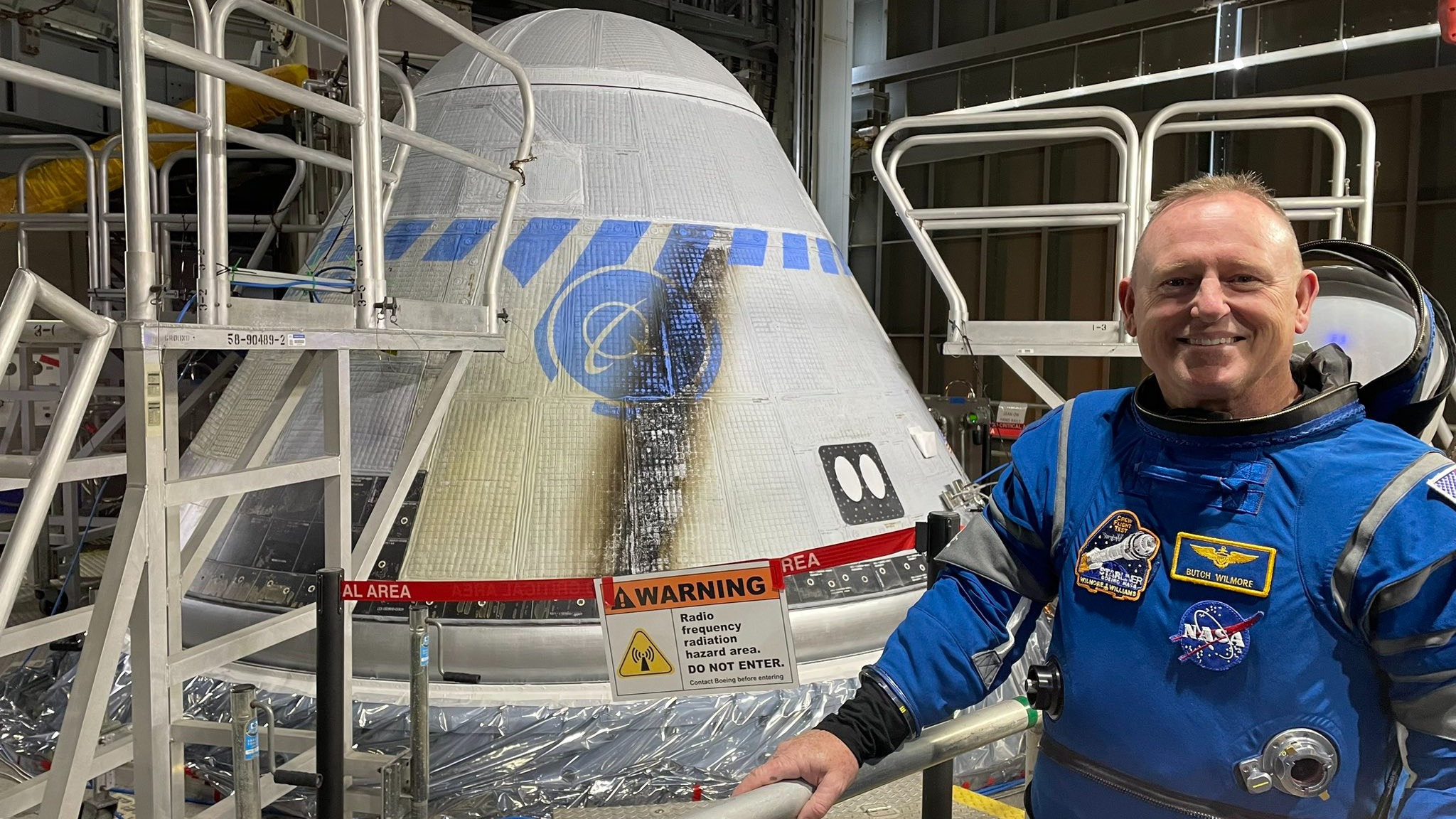 Een astronaut glimlachend naast een kegelvormig ruimtevaartuig.  Het ruimtevaartuig bevindt zich in de lanceerbrug en is omgeven door trappen.  Een brandvlek geeft de zijkant van het ruimtevaartuig aan