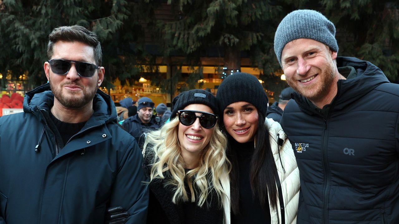 Prince Harry smiling next to Meghan Markle, Michael Buble and his wife standing outside in Whistler, Canada, all wearing winter coats and hats