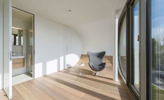 A grey chair with a white side table in a room in a residential home with floor to ceiling windows, wooden floors, white walls and ceilings.