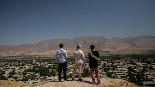 Three people stand on a sandy hilltop, overlooking a town with mountains in the distance.