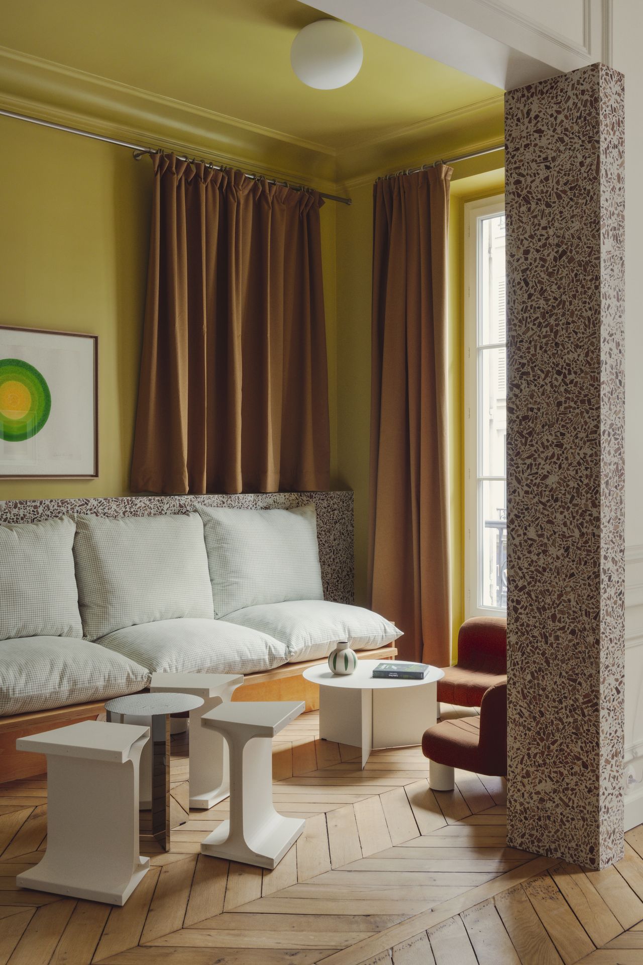 A modern living space with yellow walls, floor to ceiling burnt orange curtains and a white cushioned sofa against the far wall. There are various coffee tables in front of the seating area and two small chairs in front of the window.