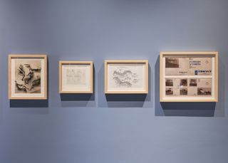 On a light blue wall are four wooden frame showing Perriand's sketches of her tubular steel chairs as well as a brochure from furniture manufacturer Thonet