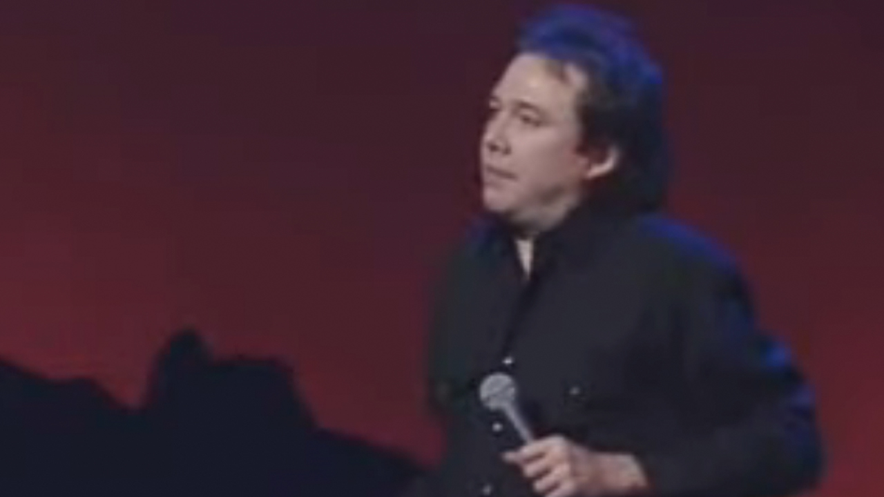 Bill Hicks in all black in front of a red background, holding a microphone