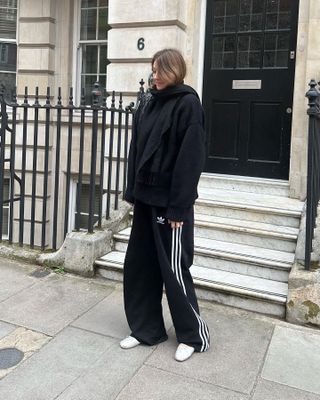 Fashion influencer Camille Charriere poses on the street in an all-black outfit with a scarf, jacket, Adidas sweatpants and white Miu Miu ballet flats