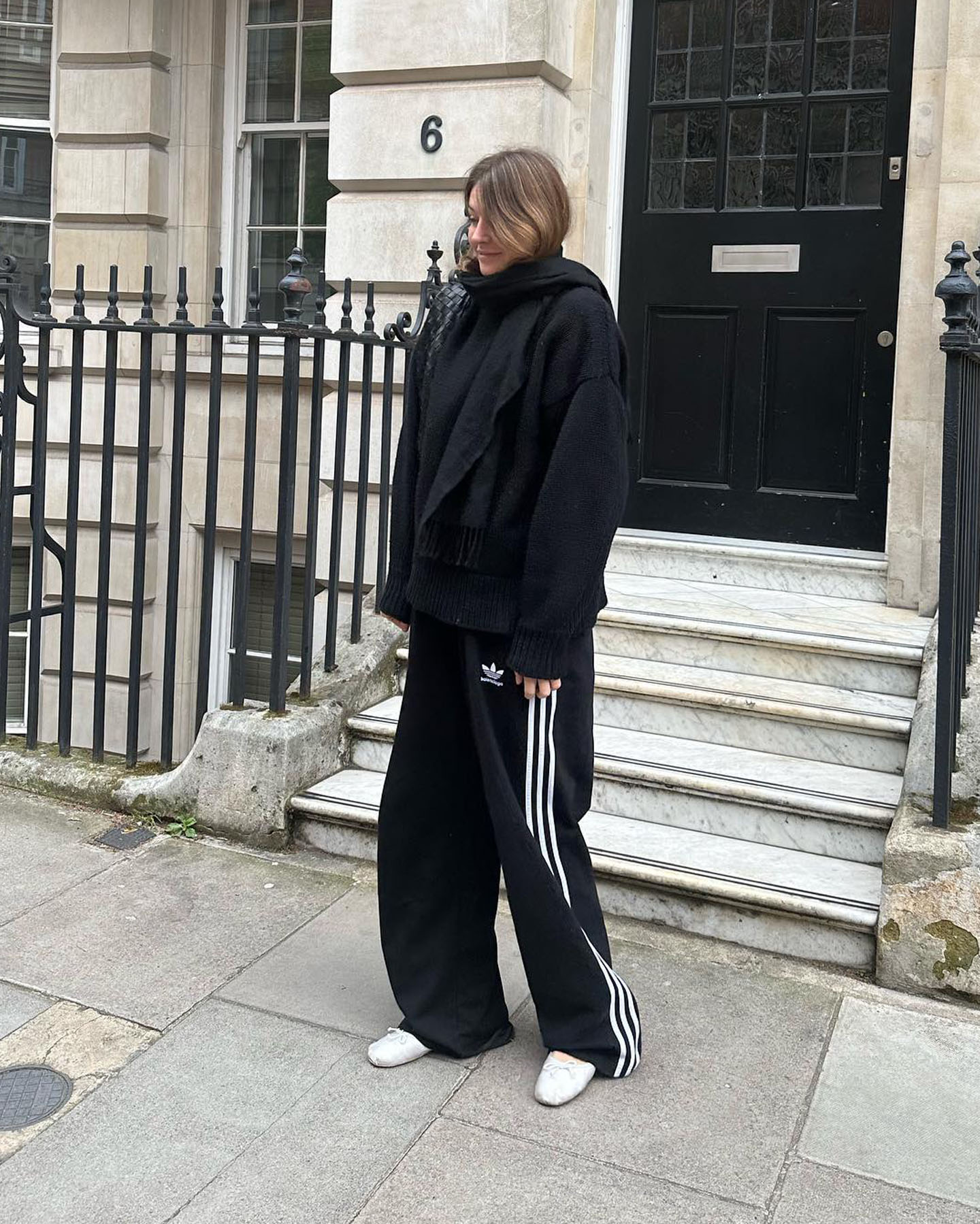 fashion influencer Camille Charriere poses on the street in an all-black outfit with a scarf, coat, Adidas track pants, and white Miu Miu ballet flats