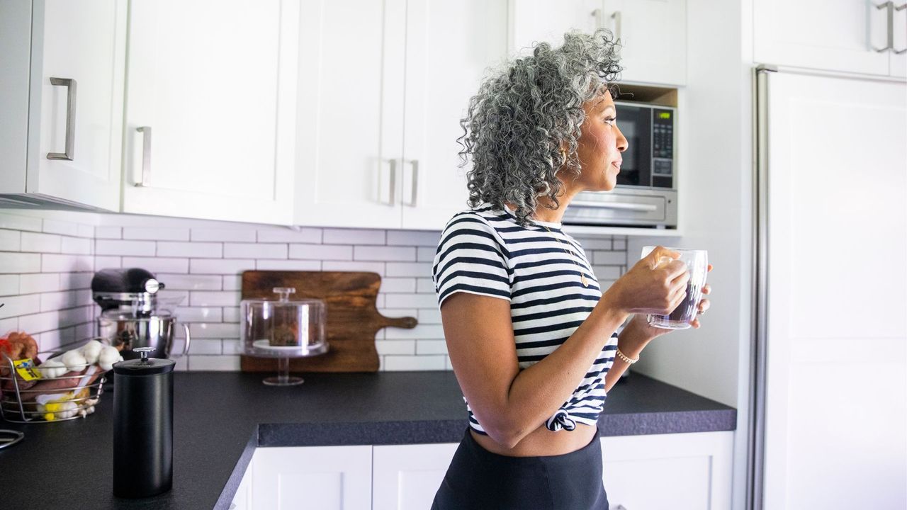 Woman wearing top and leggings standing in kitchen, holding mug of coffee, representing what to do after a bad night&#039;s sleep