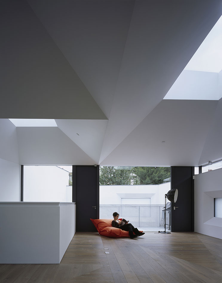 A large open space in a residential home, with wooden floors, white ceilings and white walls. A lady is sitting on a huge orange cushion o nthe floor nest to a black floor lamp