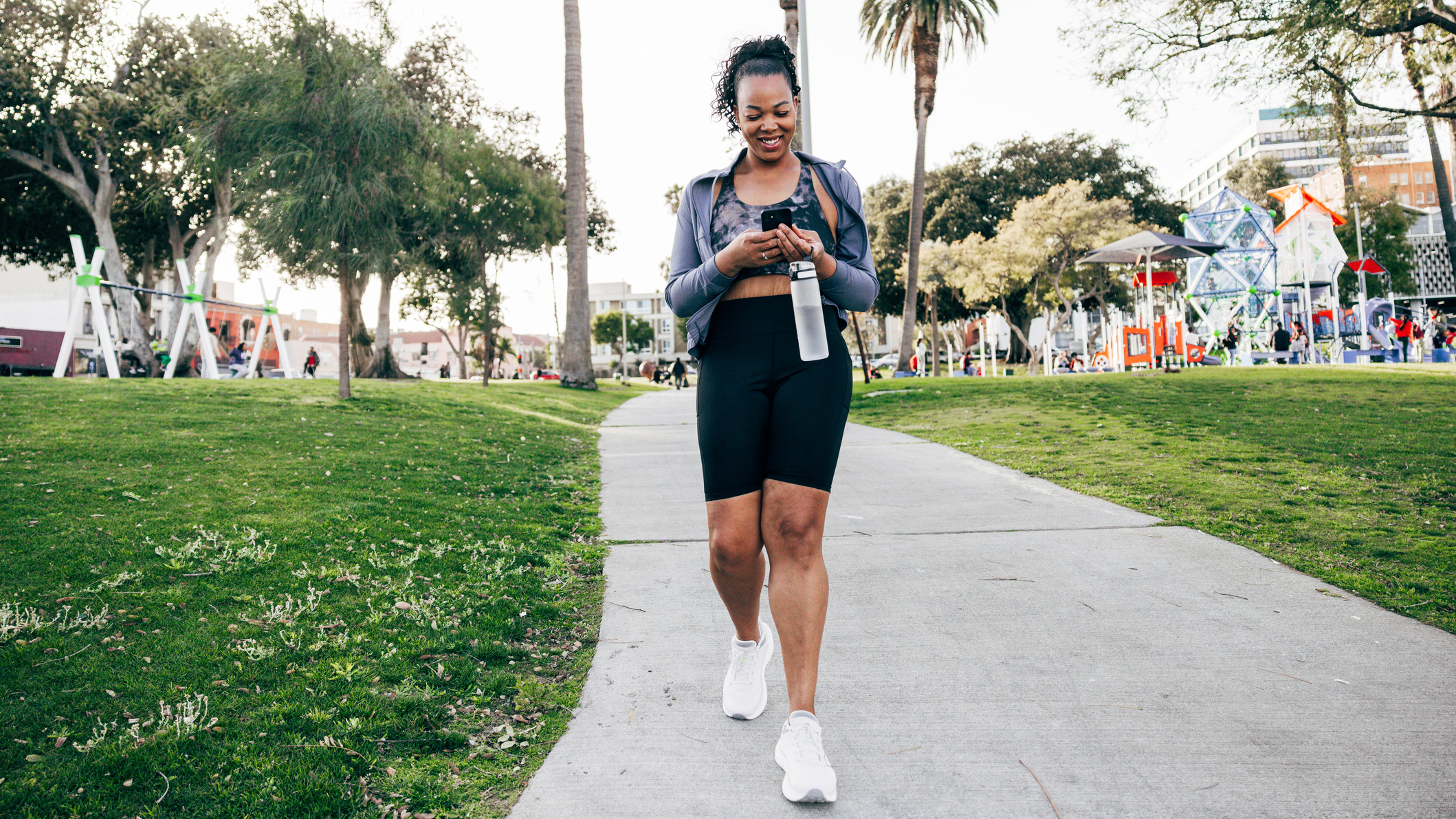 Smiling woman walking along path carrying water bottle and looking at smartphone
