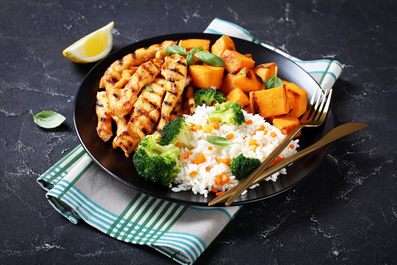 A plate of grilled chicken, rice, broccoli and cubed sweet potatoes sat on a tea towle on a black marble backdrop.