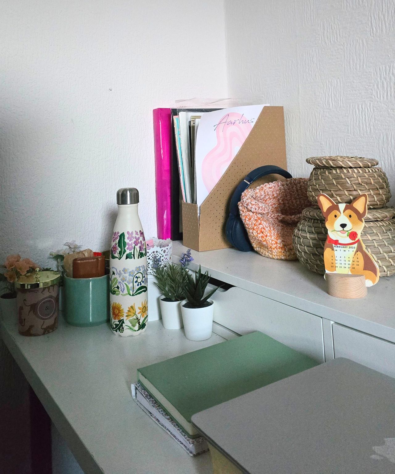 A white two-tiered desk with circular woven tubs, a magazine filer, candles, a water bottle, a green notepad, and a silver laptop on top of it