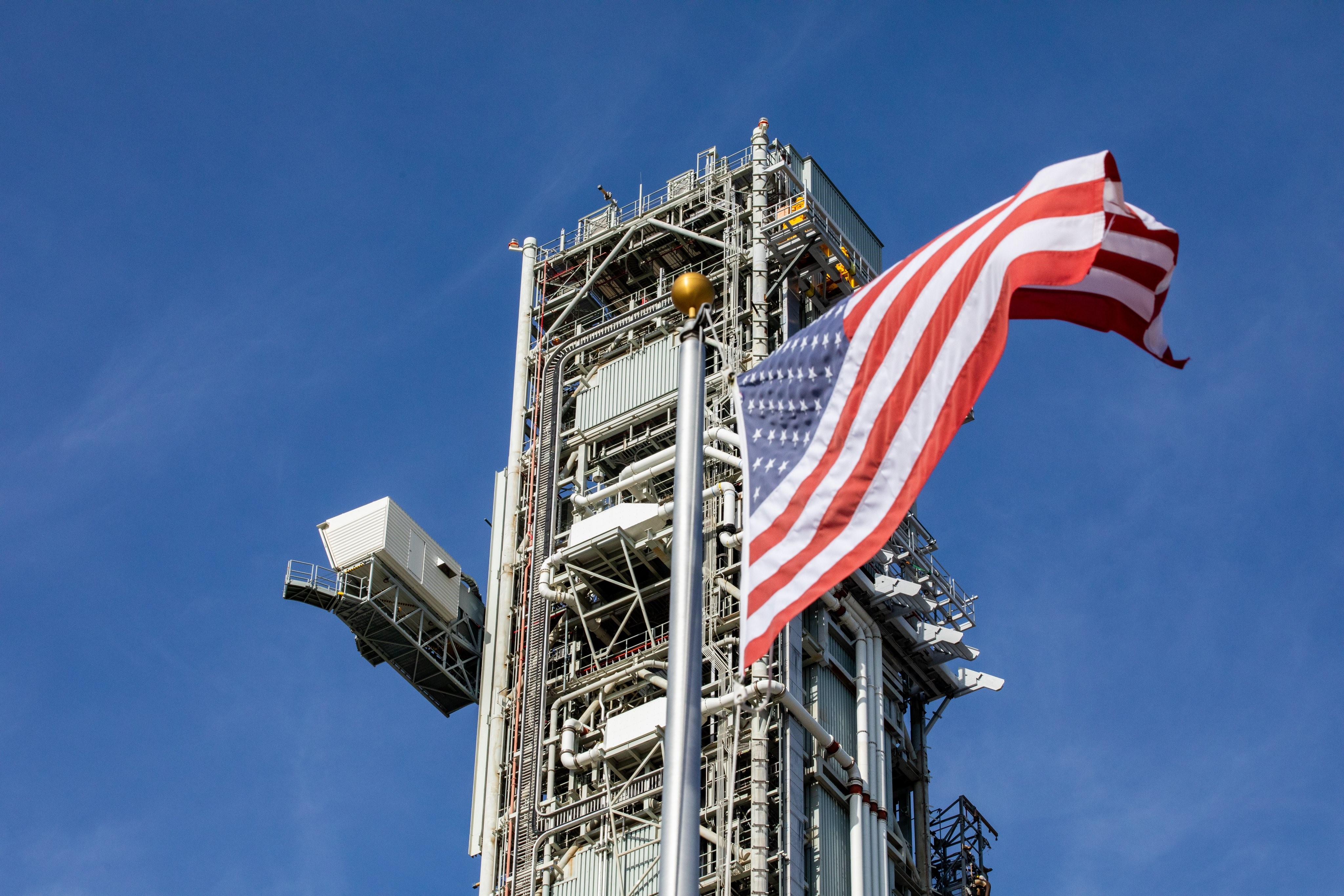 Mobile launcher 1 is seen in the background while an American flag is flying in the wind in the foreground. The crawler is carrying the mobile launcher 1 from the park site to Launch Pad 39B. The sky is partly cloudy and blue. Mobile launcher 1 has the crew access arm showing to the left and many intricate pieces are seen throughout.