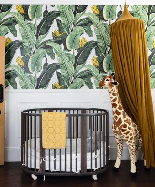 nursery with green botanical wallpaper, black crib with large giraffe soft toy and mustard yellow ceiling canopy