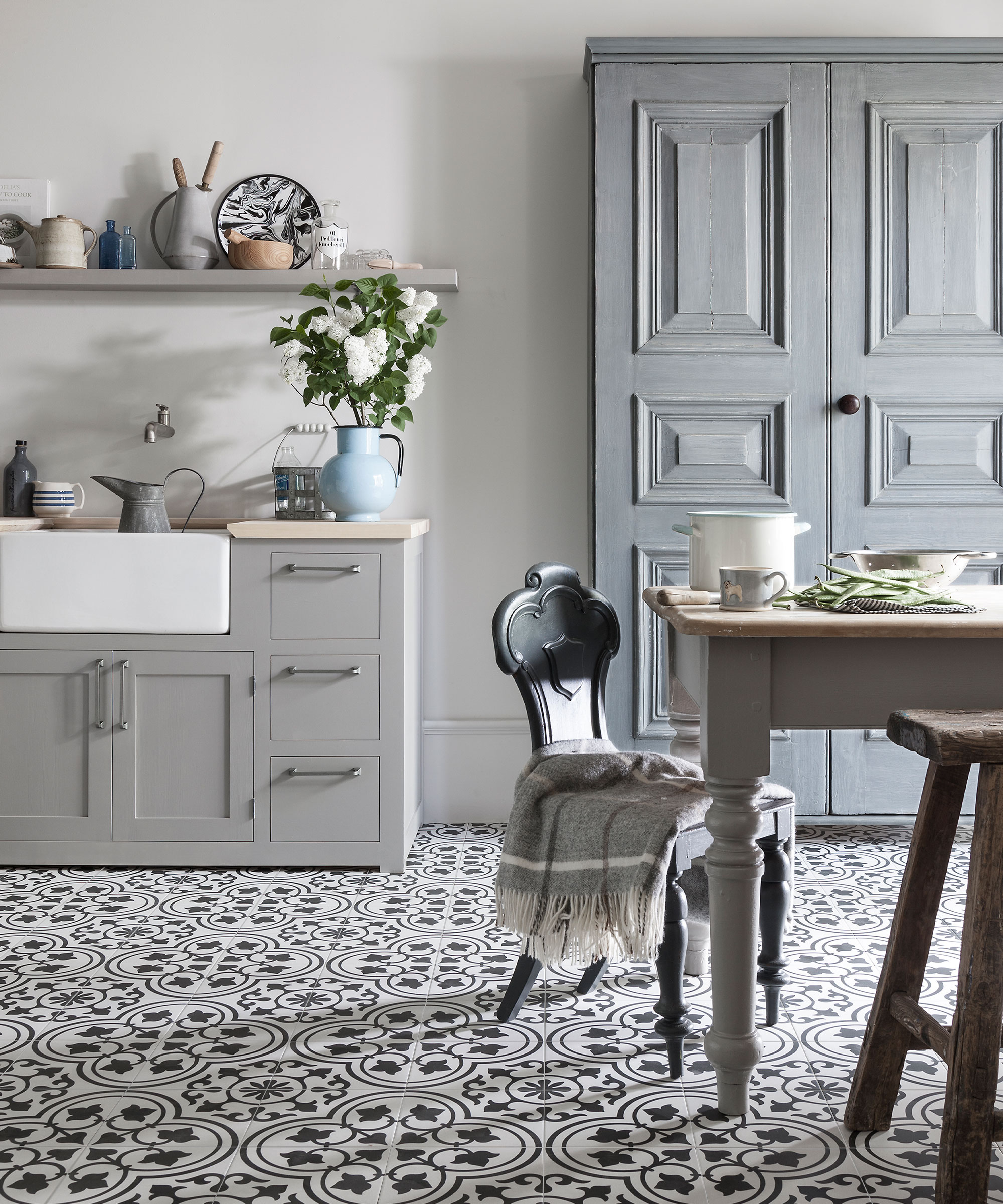 Dining-kitchen space with cuban, black and white floor tiles, traditional wooden dining table with gray painted frame, gray painted cabinet and kitchen unit, gray shelf decorated with ornaments and kitchen accessories