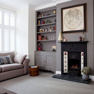 A grey living room with black fireplace, grey sofa, grey carpet, book shelves and white window shutters