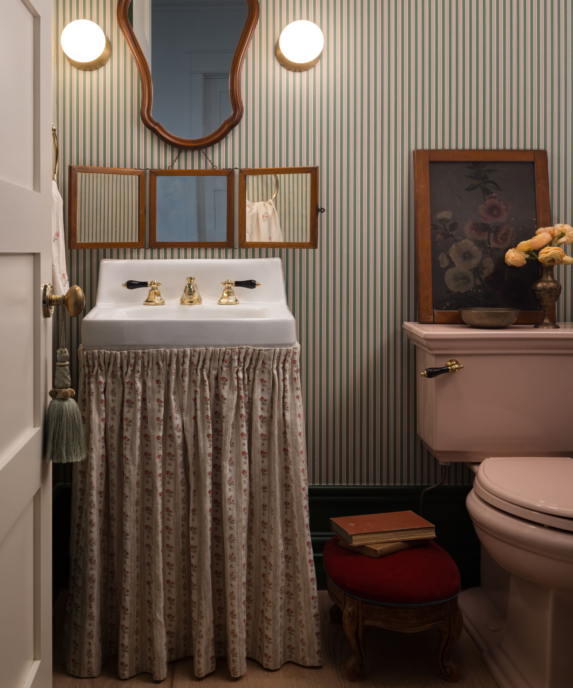 a compact powder room with stripe wallpaper, small skirted sink and a pink toilet styled with vintage mirror, art and accessories