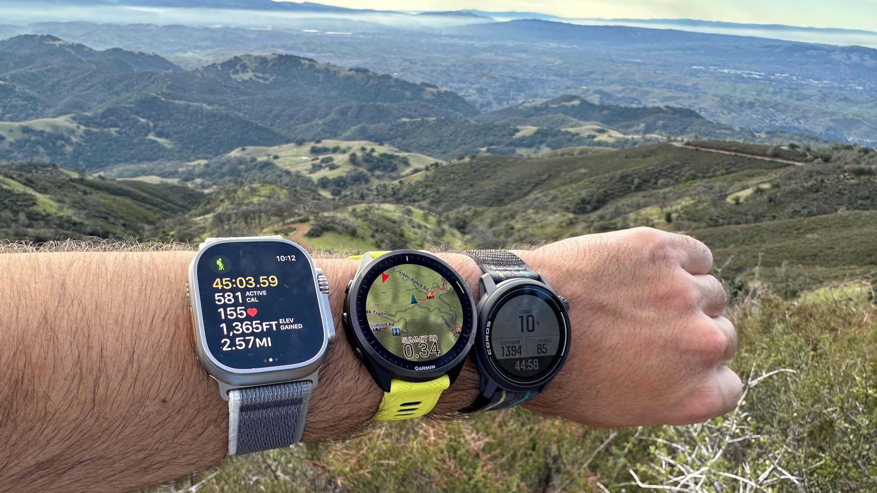 The Apple Watch Ultra 2, Garmin Forerunner 965, and COROS PACE 3 are all worn on one wrist on the Mount Diablo Summit Trail, overlooking the valley below.