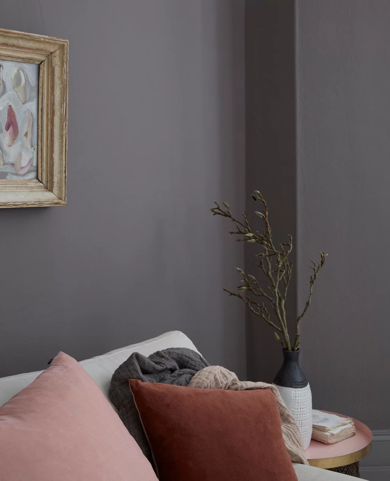 brown living room with artwork in gold frame, white sofa with pink and orange pillows, and a side table styled with a book and a vase of stems