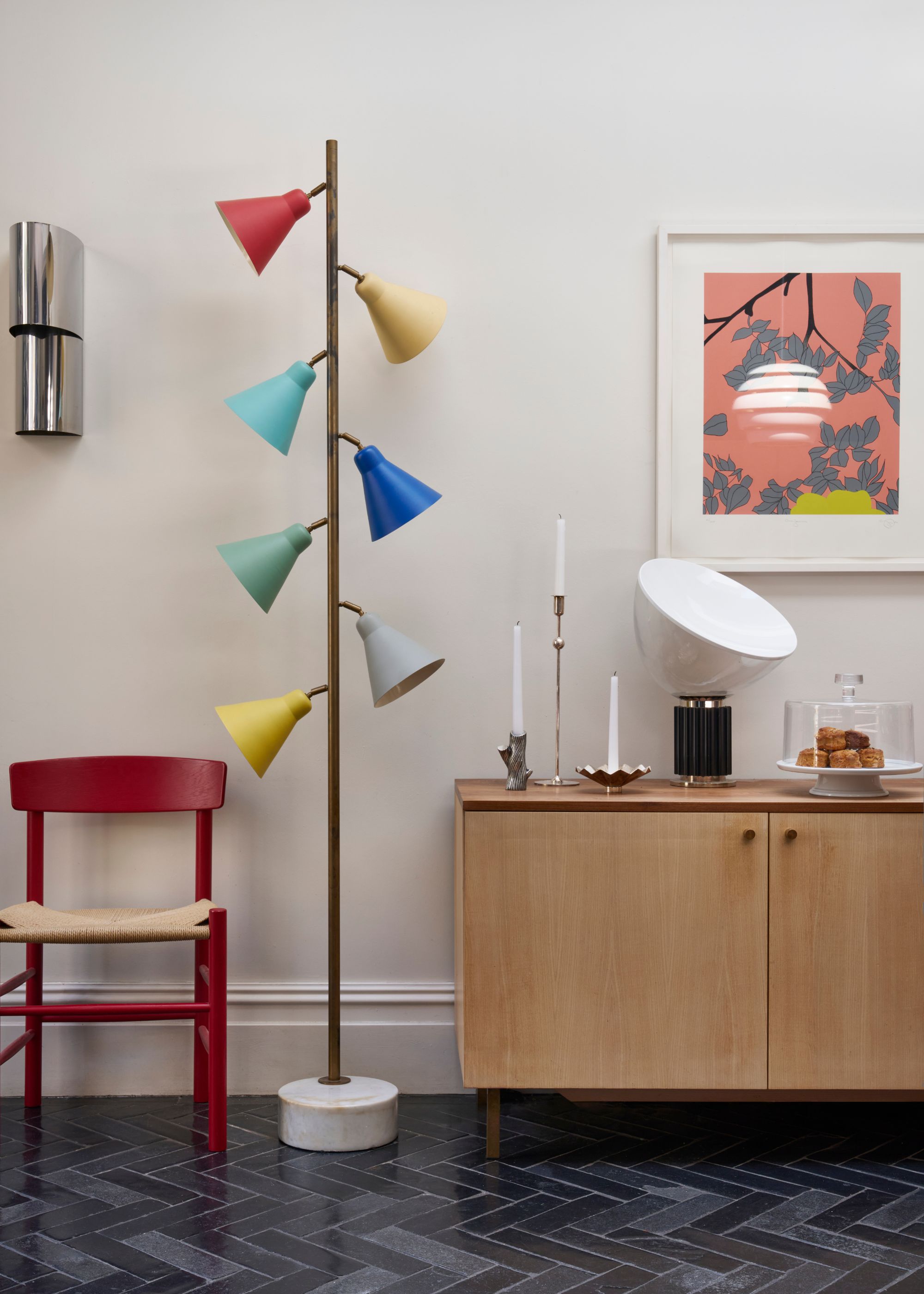 An area of the kitchen with a storage nook as well as a colorful floor lamp next to the kitchen console. On the console is a tray with baked goods, a candle and a table lamp. Above the console is a framed piece of art. There is also a red painted chair placed beside the floor lamp.