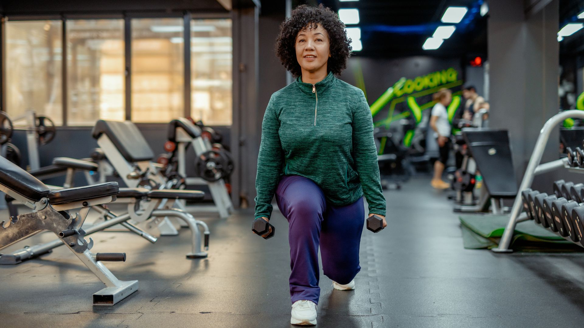 Woman doing reverse lunge, one of the best exercises for 60 and over in the gym holding dumbbells and wearing activewear