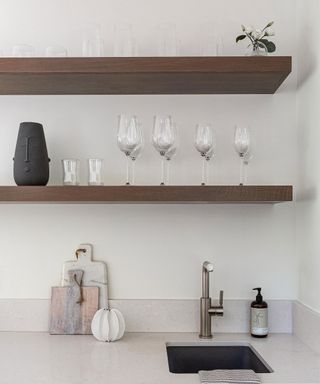 Neutral, relaxed kitchen space with two dark wood shelves, decorated with glassware, ornaments, white painted walls, light gray stone countertop