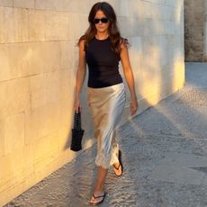 British influencer Kim Turkington walking during golden hour in Palma De Mallorca, Spain wearing a chic summer outfit with a black sunglasses, a black sleeveless top, silk ivory skirt with lace hem, a black beaded hand bag, and black flat flip-flop sandals.
