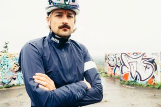 A white man with his arms crossed in a blue waterproof cycling jacket looks directly into the camera