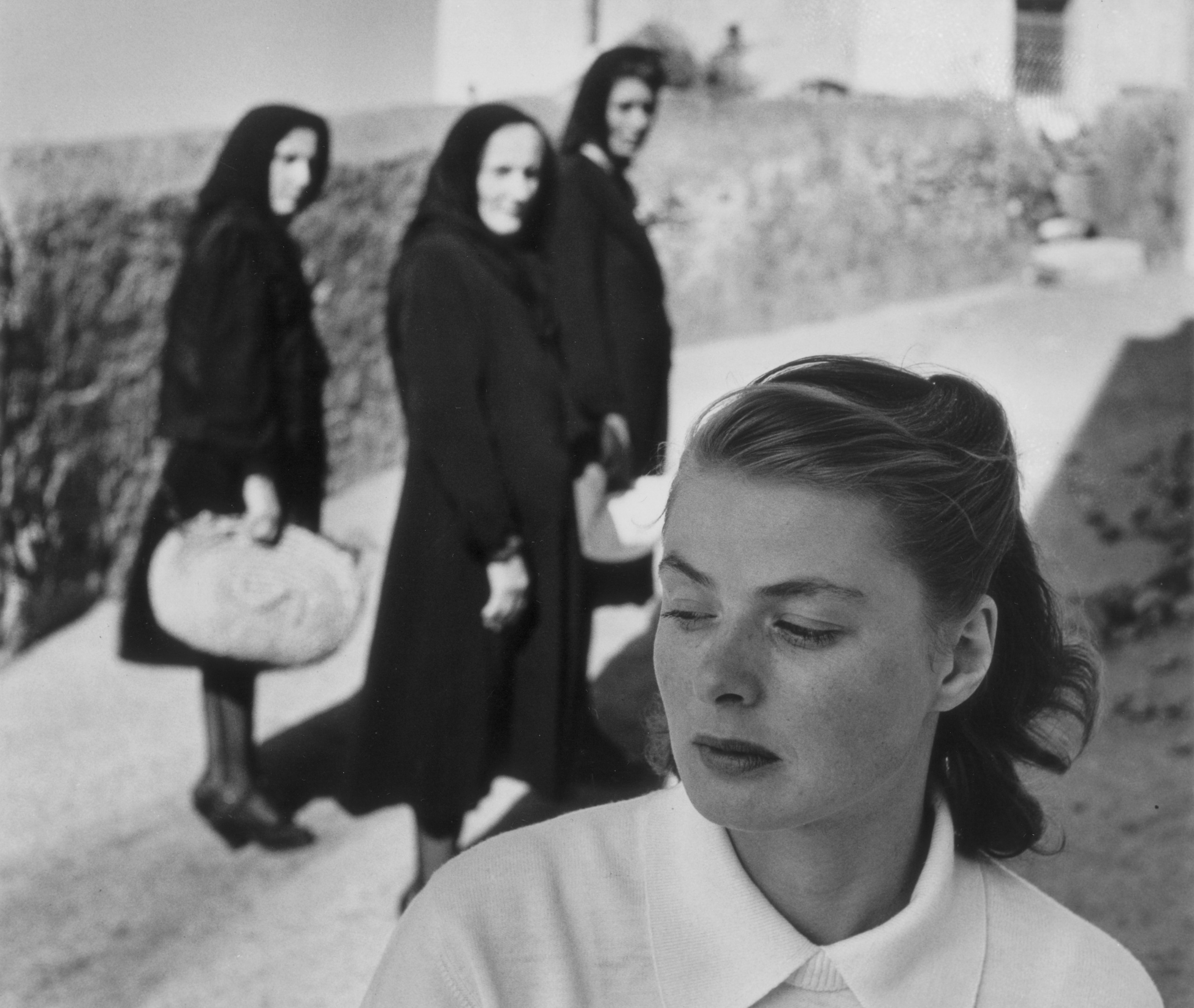 Ingrid Bergman looks down and to the side in the foreground, while three women in dark, hooded robes carrying bundles pass behind her along a sunlit stone wall.