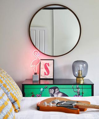 Bedroom with round mirror on white wall, green monkey chest of drawers and electric guitar on bed. S artwork print. Recently renovated Victorian villa in North London, home of Diana Potts and her family.