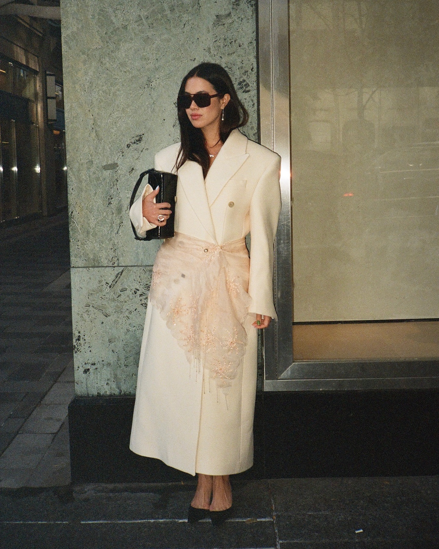 Woman wears a double breasted white coat with dark sunglasses, black heels and a printed silk scarf tied around her waist.