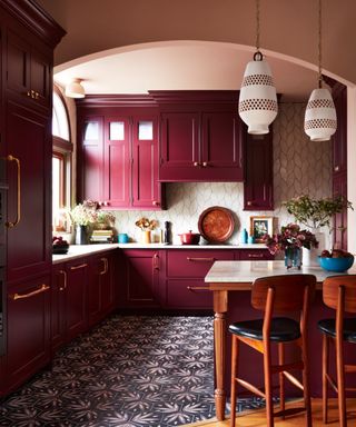 burgundy kitchen with burgundy cabinets, blue patterned floor tiles, kitchen island, pearlescent tiled wall/backsplash, plaster pink walls and ceiling, white pendant lights, brass hardware