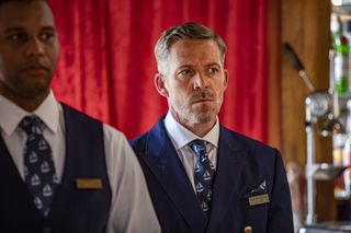 Death In Paradise: Marlon Collins (Sean Maguire) stands in his uniform at the Saint Marie Yacht Club, wearing a badge that says "Manager". He has a sombre look on his face.