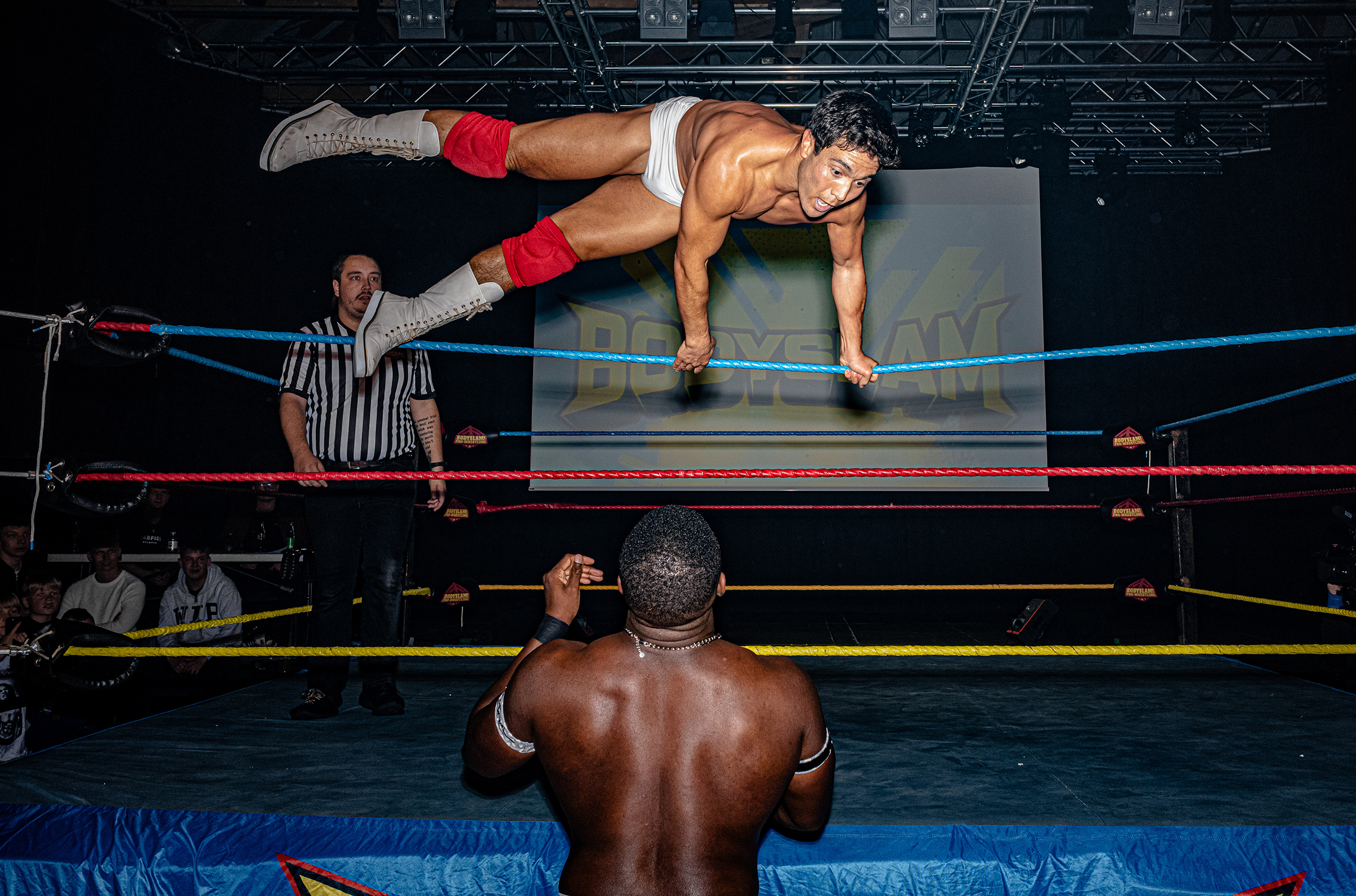 A wrestler in white trunks and red boots leaps mid-air inside a wrestling ring. Another wrestler stands below, surprised. A referee observes the scene