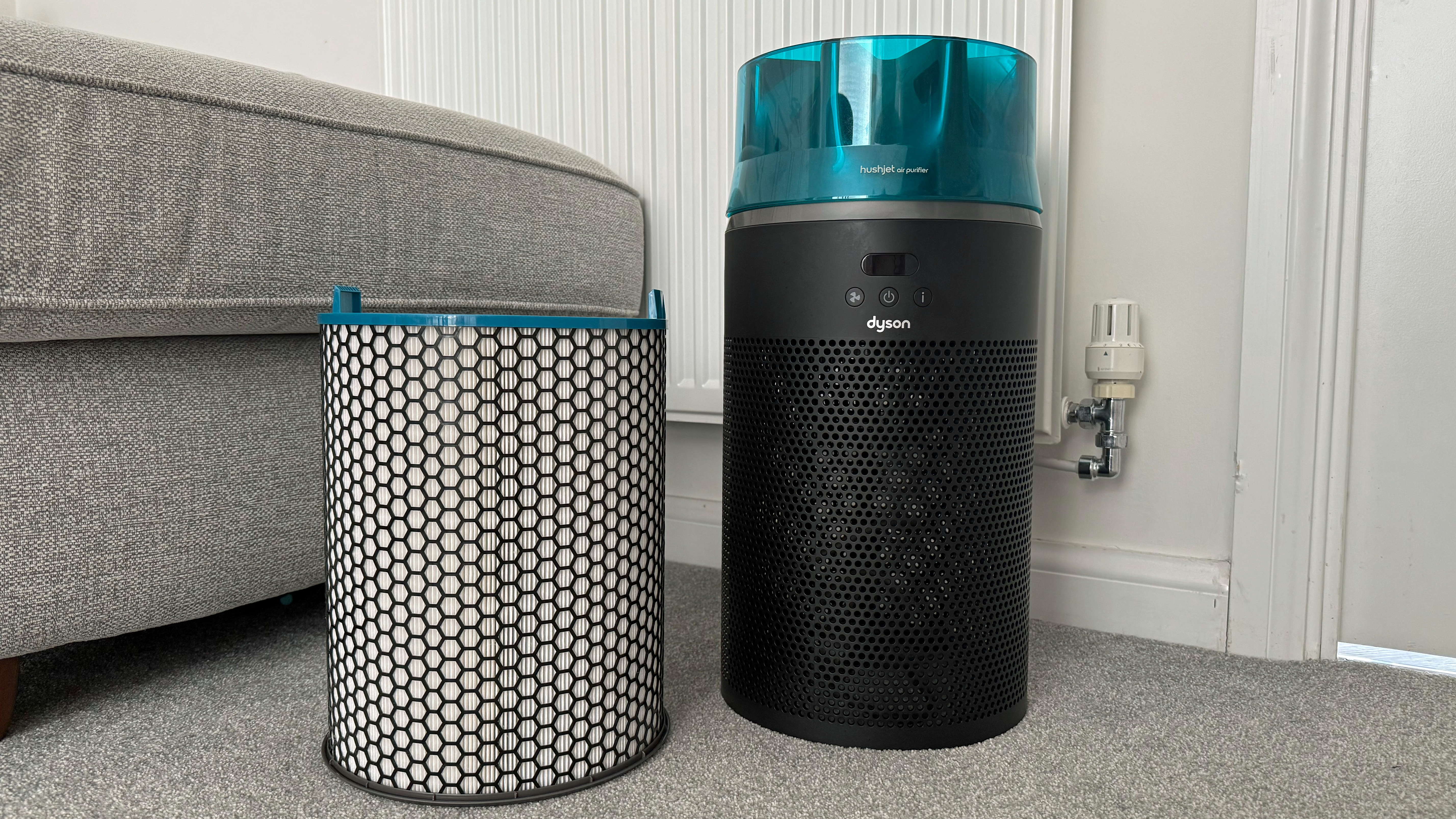 Dyson HushJet air purifier positioned beside its cylindrical replacement filter on a carpeted floor near a sofa and radiator