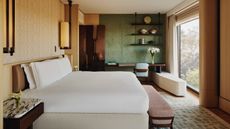 Image of a mid-century style hotel room with a large bed in the middle that is dressed in white linens. The walls are beige, and the back wall is green. There is wooden furniture around the room and large sconce lighting on either side of the bed.