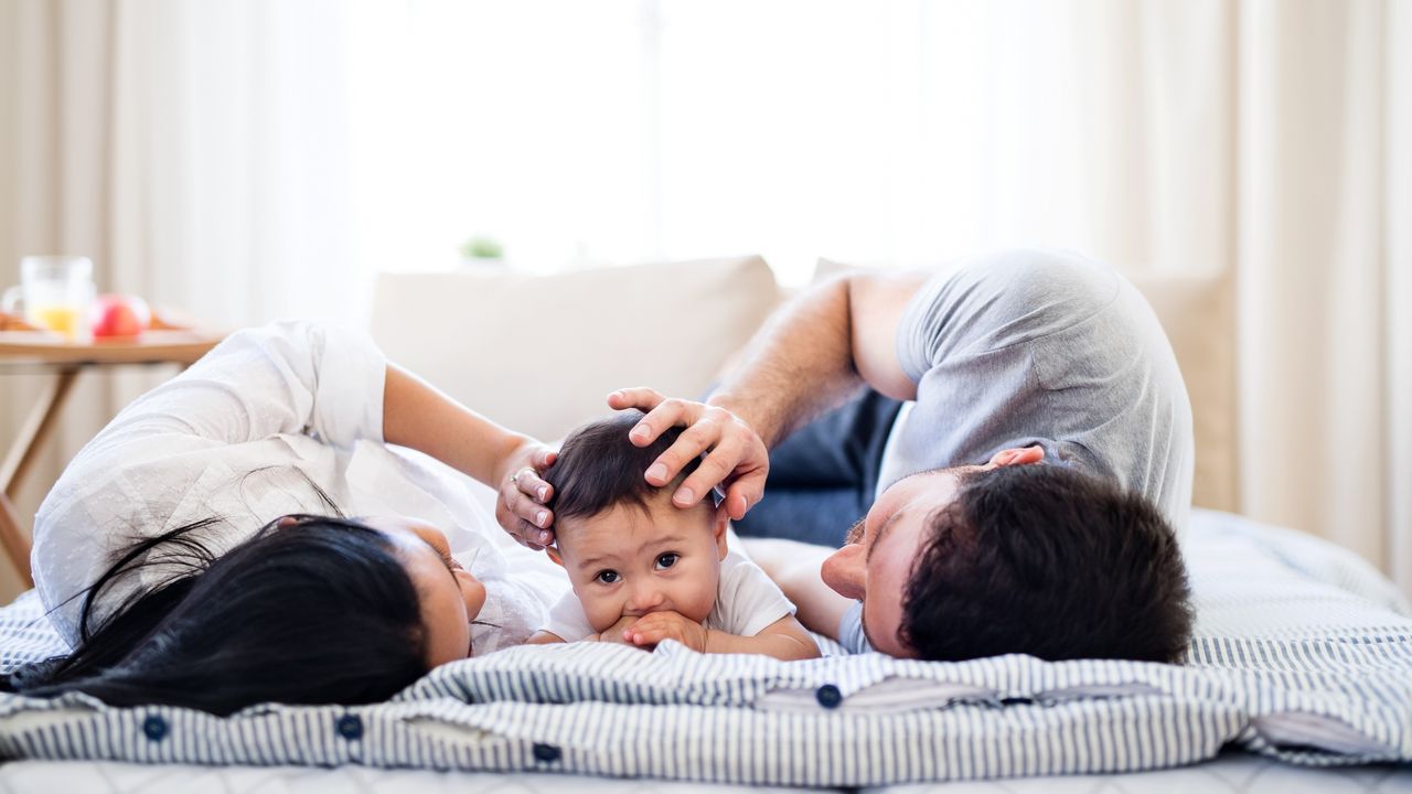 A young mom and dad lie on the bed with their toddler.