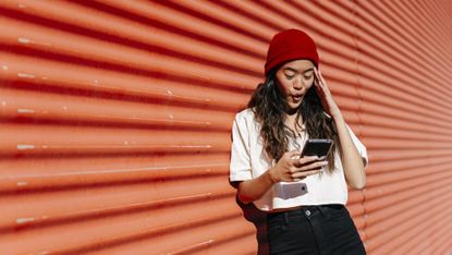 woman texting with surprised look on her face while outside against a red wall