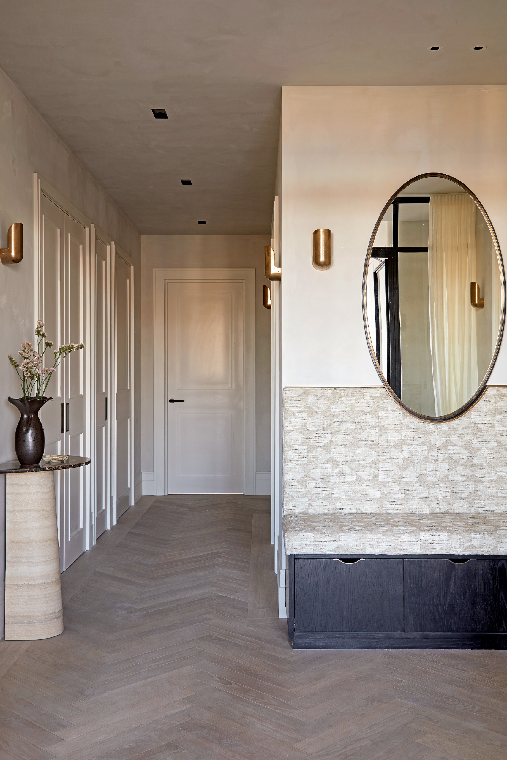Hallway with built-in bench in black stained wood with beige patterned upholstered seat and back and an oval mirror cutting into the back